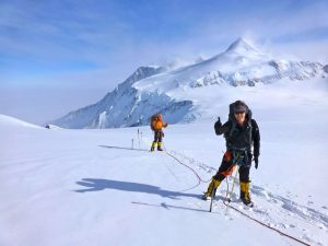 Two climbers in winter gear, supported by Antarctic Logistics and Expeditions, are roped together on a snowy mountain slope in Antarctica. The lead climber gives a thumbs-up and smiles, with a snow-covered peak rising in the background.