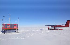 A red and blue research station with wind turbines stands on the snowy landscape of Antarctica, with a red twin-engine plane and people nearby under a clear blue sky—showcasing Antarctic Logistics and Expeditions at work.