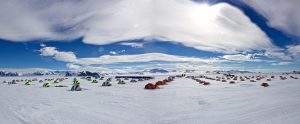 A large field of colorful tents, set up by Antarctic Logistics and Expeditions, brightens a snowy expanse under a sunny, cloudy sky, with Antarctica’s majestic mountains visible in the distant background.