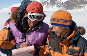Two people in winter gear stand on snow near snowy mountains, one in white ski goggles and a purple vest pointing at a document. The other, in an orange hat and jacket, looks on—an Antarctic Logistics and Expeditions team at the South Pole.