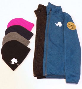 Four beanies (pink, navy, gray, black) and two jackets (black, blue) are arranged on a white surface. All items feature a white Antarctica logo, celebrating Antarctic Logistics and Expeditions at the South Pole. The blue jacket has a circular sleeve patch.