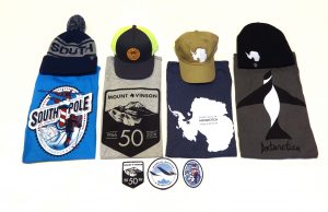 Three hats, three t-shirts, and three patches with Antarctic themes are arranged on a white background. The shirts, hats, and patches feature maps, logos, and graphics related to Antarctic Logistics and Expeditions and the South Pole.