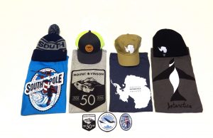 A display of three t-shirts, three hats, and four patches or stickers with South Pole and Antarctica themes—each item features related graphics or Antarctic Logistics and Expeditions logos, all arranged neatly on a white background.
