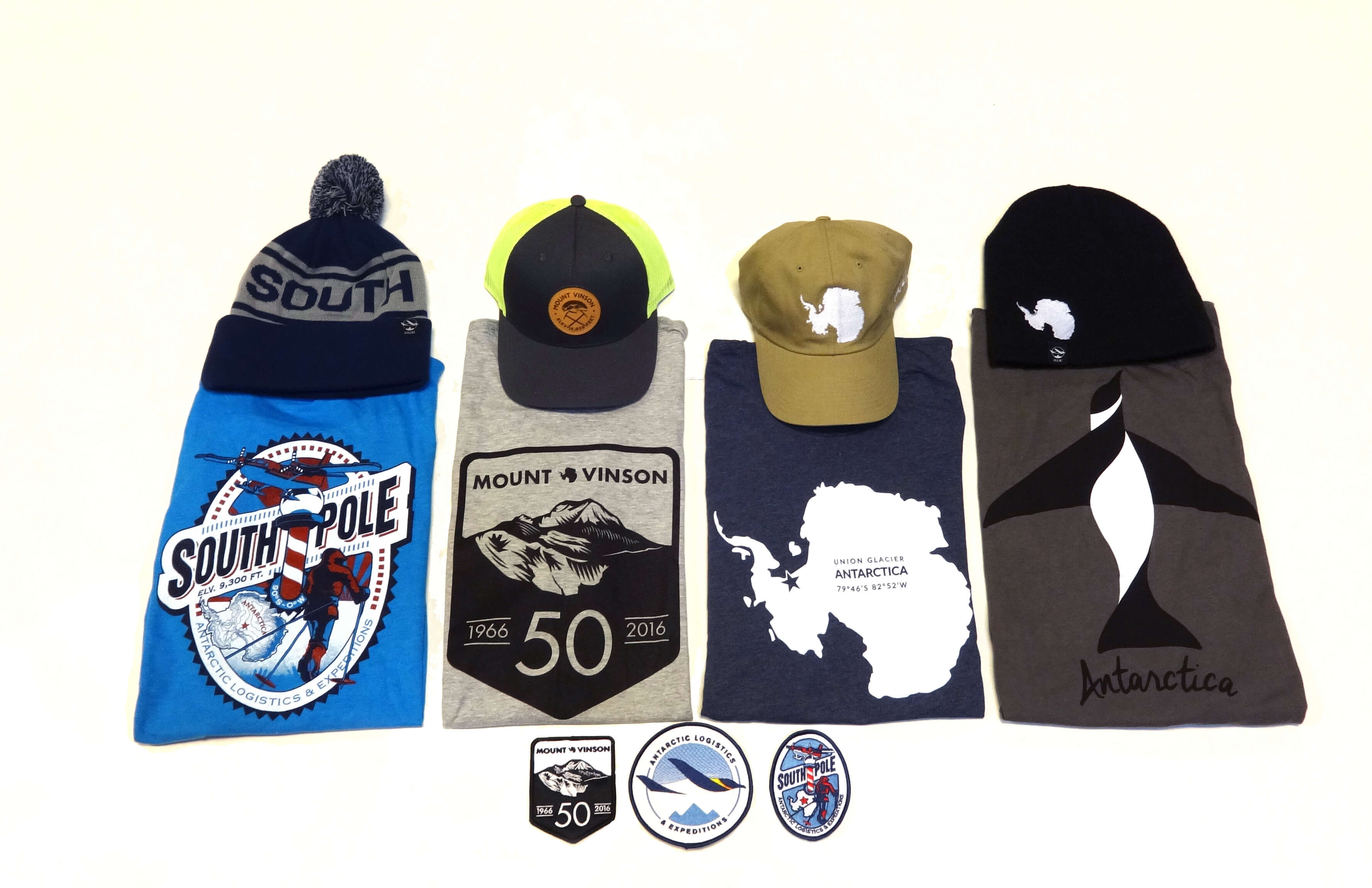 A display of three t-shirts, three hats, and four patches or stickers with South Pole and Antarctica themes—each item features related graphics or Antarctic Logistics and Expeditions logos, all arranged neatly on a white background.