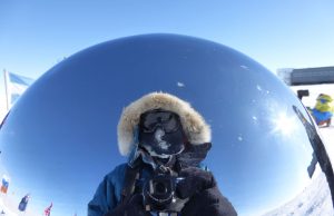 A person in cold-weather gear takes a photo of their reflection in a shiny, metallic sphere at the South Pole, with snow, flags, and blue sky visible in the background—an iconic scene from Antarctica.