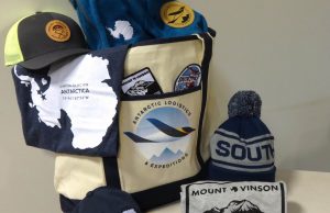 A tote bag with an Antarctic Logistics and Expeditions logo is surrounded by Antarctica-themed gear, including t-shirts, a trucker hat, a beanie reading “SOUTH,” stickers, and a Mount Vinson patch inspired by the South Pole.