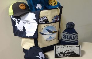 A canvas bag with an Antarctic Logistics and Expeditions logo holds stickers, a blue towel, and a shirt with an Antarctica outline. Caps, a beanie labeled SOUTH, and a Mount Vinson tee are arranged around the bag—perfect for South Pole enthusiasts.