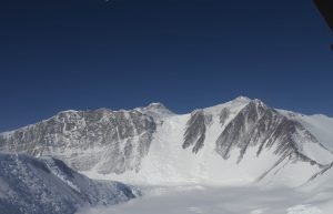 Snow-covered mountain peaks under a deep blue sky, with steep rocky slopes and a glacier in the foreground capture the remote, bright beauty of Antarctica—evoking the spirit of Antarctic Logistics and Expeditions.