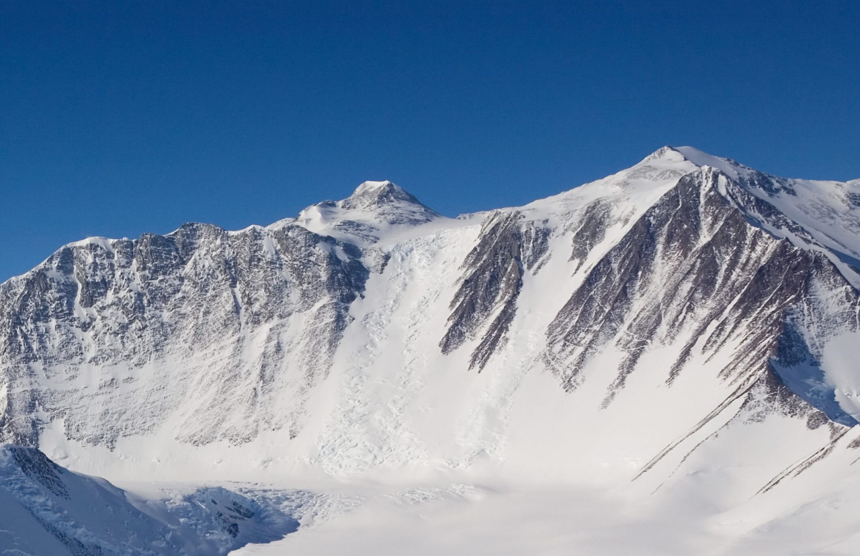 Snow-covered mountains with steep, jagged peaks rise against a clear blue sky in Antarctica. The slopes are streaked with rocky outcrops and patches of ice, and a glacier descends between the peaks into the valley below.