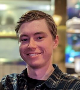 A young man with light skin and short, light brown hair smiles at the camera. He is wearing a dark plaid shirt, and the softly blurred background with colorful lights evokes the adventure of Antarctic Logistics and Expeditions in Antarctica.