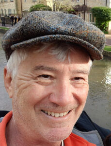A smiling older man with gray hair and a flat cap stands in front of a canal, looking as if he's ready to share stories from Antarctic Logistics and Expeditions or adventures at the South Pole. Stone buildings and greenery complete the charming background.