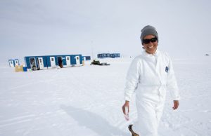 A person wearing a white snowsuit, gray beanie, and sunglasses smiles while standing on snow near blue buildings in remote Antarctica under an overcast sky, hinting at an adventure with Antarctic Logistics and Expeditions.