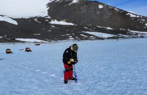 A person in winter gear drills into the icy surface of Antarctica, with snowmobiles and snowy mountains in the background under a clear blue sky—capturing the spirit of Antarctic Logistics and Expeditions.