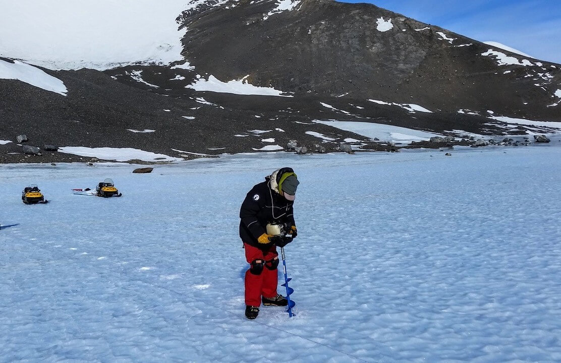 A person in winter gear drills into the icy surface of Antarctica, with snowmobiles and snowy mountains in the background under a clear blue sky—capturing the spirit of Antarctic Logistics and Expeditions.