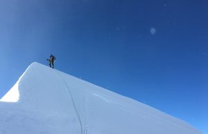 A climber stands on a sharp, snowy ridge in Antarctica under a clear blue sky, holding a rope that trails down the slope—embodying the spirit of Antarctic Logistics and Expeditions.