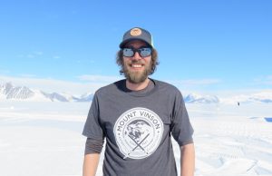 A man with a beard and sunglasses, wearing a Mount Vinson T-shirt and a cap, stands smiling in Antarctica's snowy, mountainous landscape under a bright blue sky.