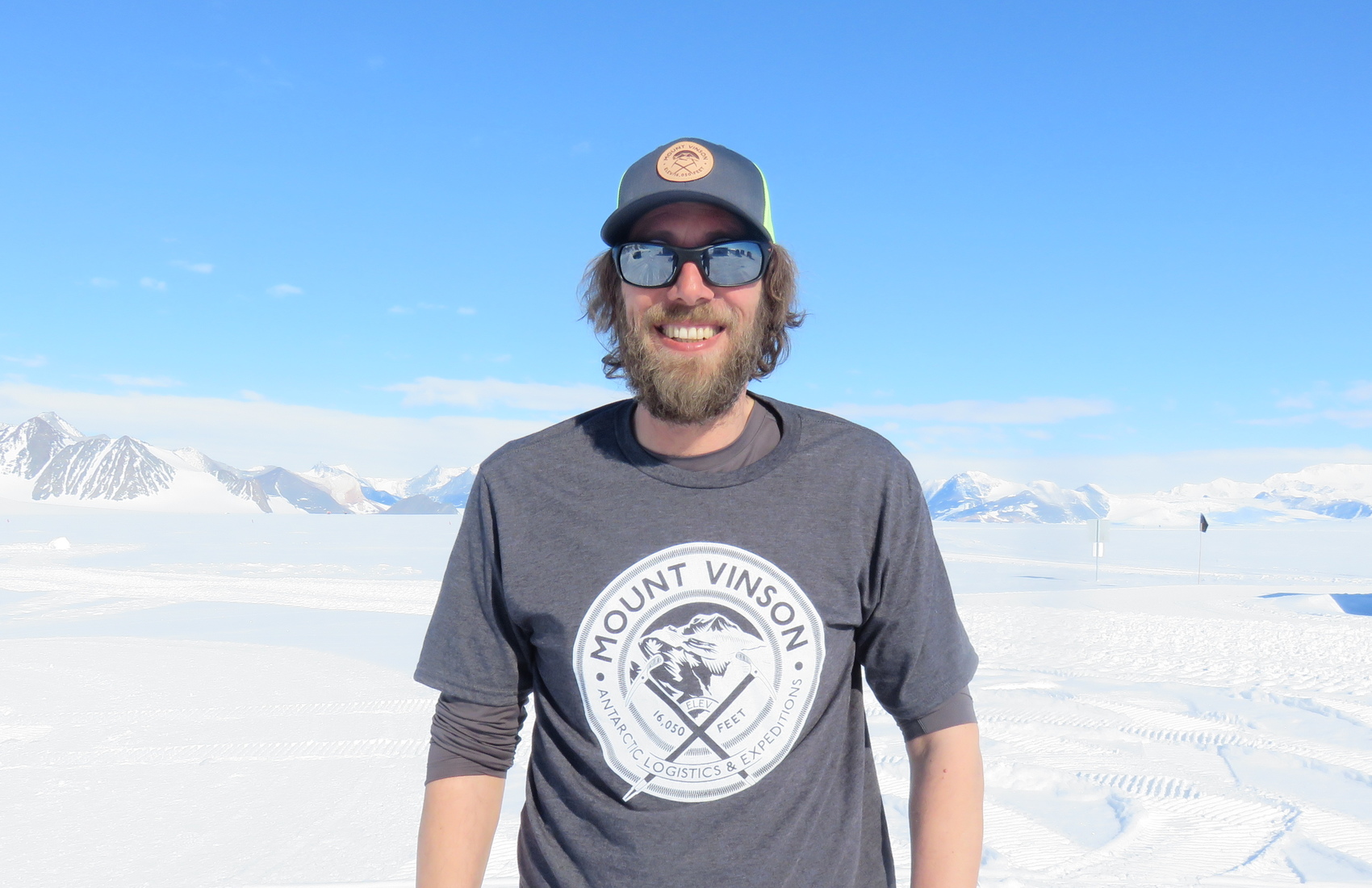 A man with a beard and sunglasses, wearing a Mount Vinson T-shirt and a cap, stands smiling in Antarctica's snowy, mountainous landscape under a bright blue sky.