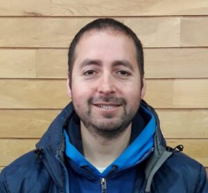 A man with short dark hair and a trimmed beard is smiling at the camera. He is wearing a blue jacket and hoodie, standing in front of a wooden panel wall, ready to share stories from his time with Antarctic Logistics and Expeditions in Antarctica.