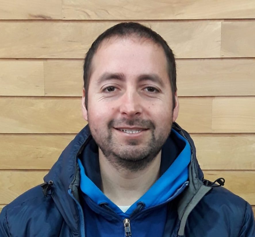 A man with short dark hair and a trimmed beard is smiling at the camera. He is wearing a blue jacket and hoodie, standing in front of a wooden panel wall, ready to share stories from his time with Antarctic Logistics and Expeditions in Antarctica.