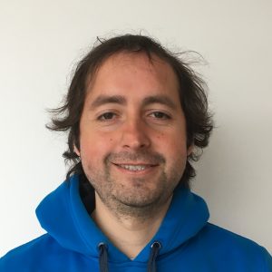A man with wavy brown hair and slight stubble smiles at the camera. He is wearing a bright blue hoodie from Antarctic Logistics and Expeditions and standing in front of a plain white background.