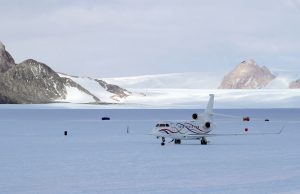 A small white jet with red and blue stripes, operated by Antarctic Logistics and Expeditions, is parked on a snowy, icy landscape in Antarctica, surrounded by mountains with patches of snow under a cloudy sky.
