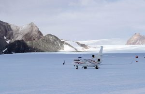 A white private jet with blue and red stripes is parked on a vast, snowy expanse in Antarctica, with rocky mountains and glaciers rising in the background under a cloudy sky—part of an Antarctic Logistics and Expeditions journey.