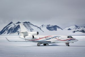 A white private jet with red and blue stripes is parked on a snowy runway, operated by Antarctic Logistics and Expeditions, with rugged, snow-covered mountains in the background under a cloudy sky near the South Pole.