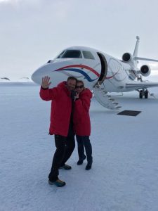 A smiling couple in red jackets stand on snow and wave, posing by a white private jet with blue and red stripes. The airplane stairs are down, and the snowy landscape hints at an Antarctic Logistics and Expeditions adventure in Antarctica.