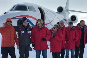 A group of seven people in red winter jackets stand smiling in front of a white private jet on snow-covered ground, ready for their Antarctica adventure. The scene appears cold and bright, with everyone dressed warmly.