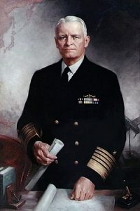 A formal painted portrait of an older man in a dark military uniform with gold stripes, holding a scroll, standing by a table with maps and books. A world map is partly visible behind him, highlighting routes to the South Pole and Antarctica.