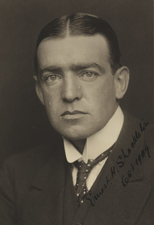 A sepia-toned portrait of a serious-looking man in a suit and tie with slicked-back hair; signed Ernest H. Shackleton Oct 1909 in black ink, evoking his legendary South Pole expeditions to Antarctica.
