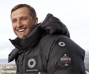 A smiling man wearing a black, insulated jacket with “Shackleton” and “Antarctic Gurkha” patches stands outdoors under a cloudy sky, evoking the spirit of Antarctic Logistics and Expeditions in a remote landscape.
