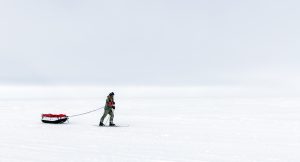 A person in winter clothing skis across a vast, snowy landscape, pulling a sled loaded with gear through the white expanse under an overcast sky on their journey toward the South Pole.