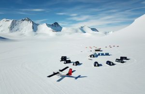 A small airplane is parked on the snowy ground near a cluster of black and white tents, part of an Antarctic Logistics and Expeditions site, surrounded by vast snowfields and distant mountains under a mostly clear South Pole sky.