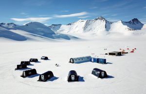 A cluster of black and white tents and buildings sit on a vast snowy landscape in Antarctica, with snow-covered mountains and blue sky in the background. Operated by Antarctic Logistics and Expeditions, more tents and people are visible in the distance.