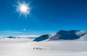 Bright sun shines over a vast, snowy Antarctica landscape with mountain peaks and deep shadows; a small encampment by Antarctic Logistics and Expeditions and tracks are visible on the snow-covered ground below the mountains.