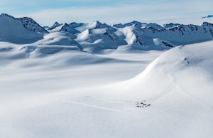 Aerial view of a snowy, mountainous Antarctic landscape with rugged peaks and vast snowfields; a small group of tents is visible on the snow, dwarfed by the South Pole wilderness and remoteness managed by Antarctic Logistics and Expeditions.