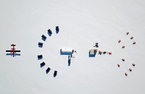Aerial view of an Antarctic snowy landscape featuring a red plane, several black vehicles in a semicircle, a central white building, and orange tents arranged in a wide arc—capturing the essence of Antarctic Logistics and Expeditions near the South Pole.