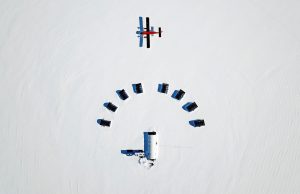 Aerial view of a small snowy encampment in Antarctica with one central building, seven smaller structures in a semicircle, and a red and black plane on the snow nearby. This remote scene showcases Antarctic Logistics and Expeditions operations.