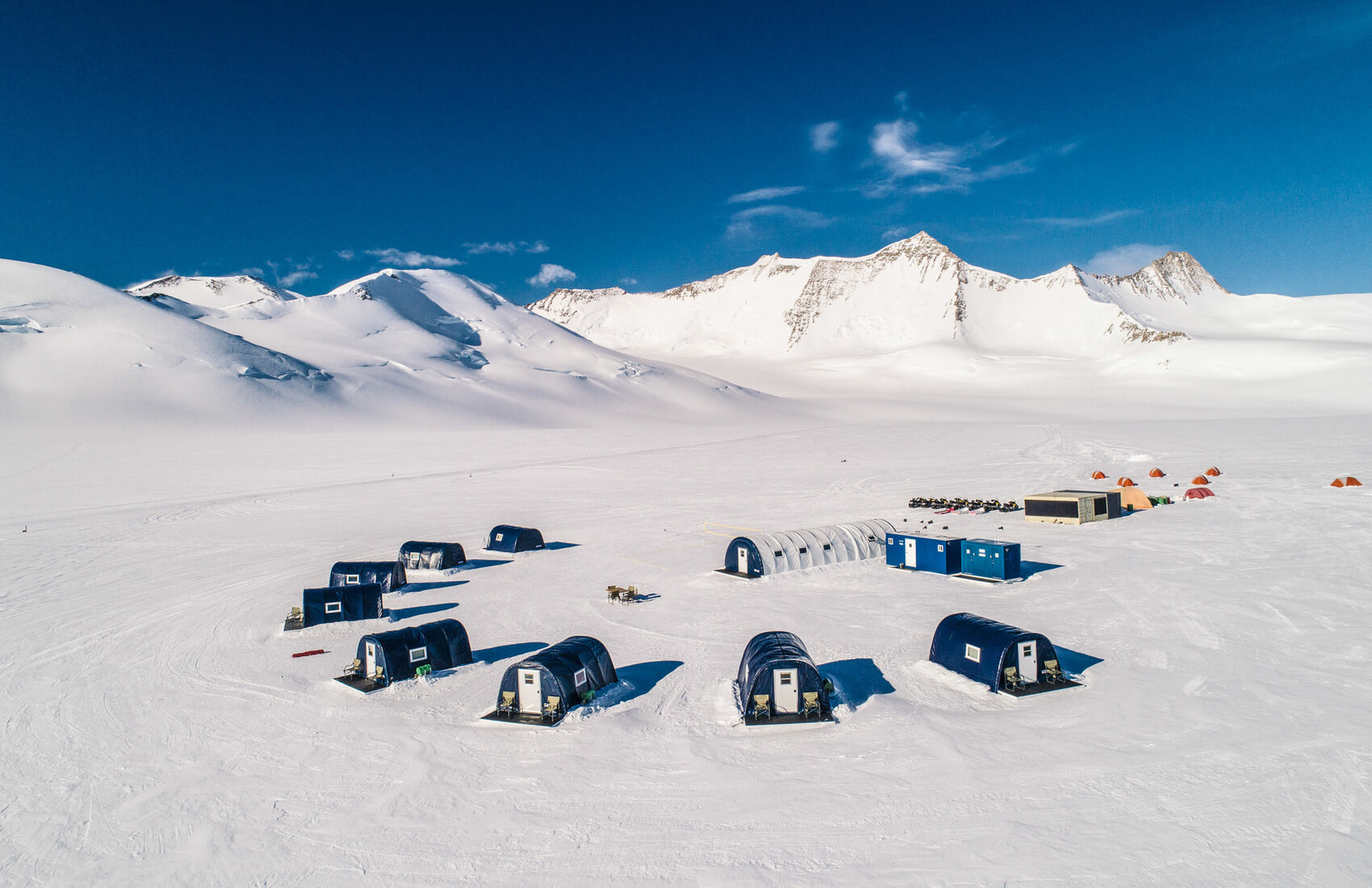 Aerial view of a remote research camp in Antarctica, with blue and white tents and small buildings set on a vast snowy plain, surrounded by snow-covered mountains under a clear blue sky.