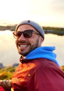 A smiling man wearing sunglasses, a gray beanie, and a red jacket over a blue hoodie sits outdoors near a calm body of water on a bright, sunny day, preparing for his upcoming Antarctic Logistics and Expeditions journey to the South Pole.