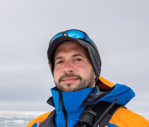 A man with a beard wears a blue and orange jacket, a gray beanie, sunglasses on his cap, and stands outdoors on a cloudy, snowy day in Antarctica near the South Pole.