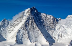 A tall, snow-covered mountain peak in Antarctica rises sharply against a clear blue sky, with wisps of clouds drifting around its summit—an iconic scene for Antarctic Logistics and Expeditions en route to the South Pole.