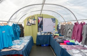 A vendor tent displays clothing and accessories on tables and hangers, including shirts, sweaters, tote bags, and hats in various colors, with an Antarctica map or Antarctic Logistics and Expeditions poster on the back wall.