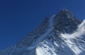 A snow-covered mountain peak rises sharply against a clear deep blue sky in Antarctica, with patches of snow and ice clinging to its rugged slopes. A small wisp of cloud drifts nearby, capturing the spirit of Antarctic Logistics and Expeditions.