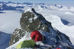 Two tents, one red and one green, are set up on a snowy mountain ridge in Antarctica with two people nearby. Rugged, snow-covered peaks and glaciers stretch across the background under a clear sky—true Antarctic Logistics and Expeditions style.