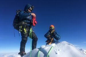 Two climbers in winter gear stand on a snowy Antarctic peak under a clear blue sky, connected by bright green ropes. One adjusts equipment while the other kneels, both wearing sunglasses and helmets—preparing for their South Pole journey.