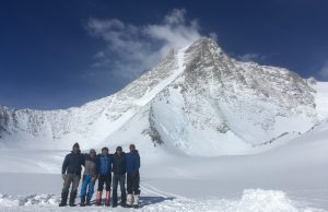 Five people dressed in winter gear stand together on snow in Antarctica, with a large, snow-covered mountain and blue sky in the background—an adventure supported by Antarctic Logistics and Expeditions.