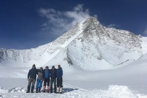 Five people in winter clothing stand together in front of a snowy mountain peak under a clear blue sky, surrounded by snow-covered ground and rugged terrain, ready for their Antarctic Logistics and Expeditions adventure.
