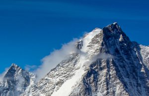 Snow-covered mountain peak in Antarctica with rugged, steep slopes rises against a vibrant blue sky, wisps of clouds clinging to its summit and ridges—a stunning scene often explored by Antarctic Logistics and Expeditions.